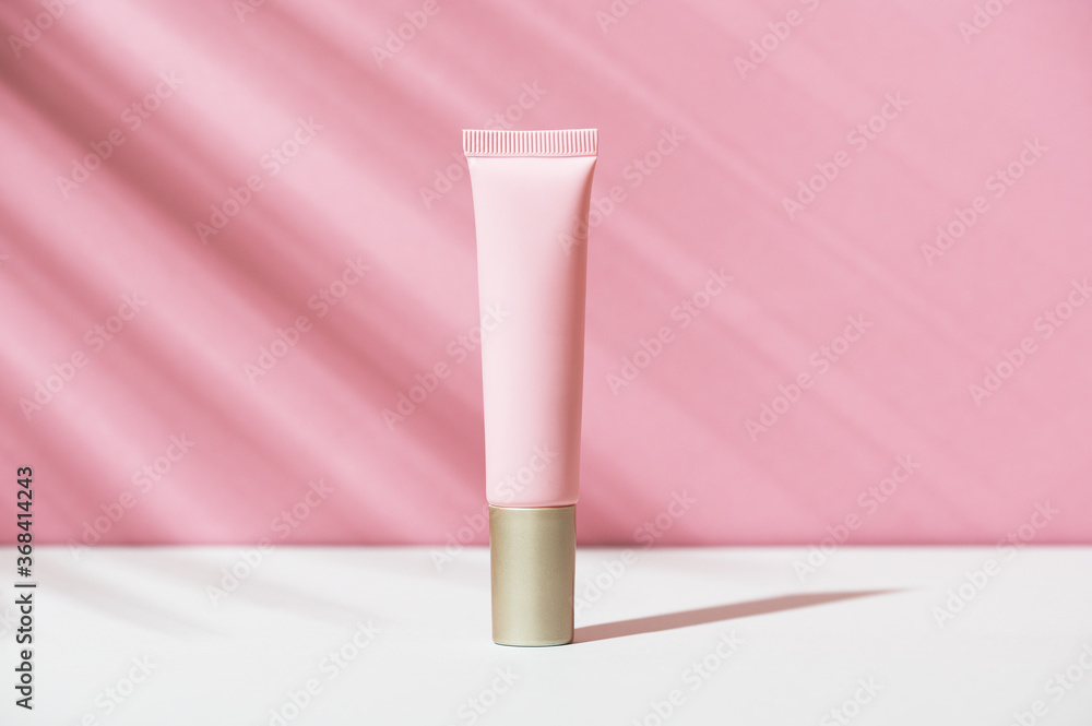Pink tube of soft organic natural eye cream with golden cap on white pink background, palm leaf shadow. Summer facial product, sun lotion. Women's hygiene cosmetic accessory for skincare. Copy space.