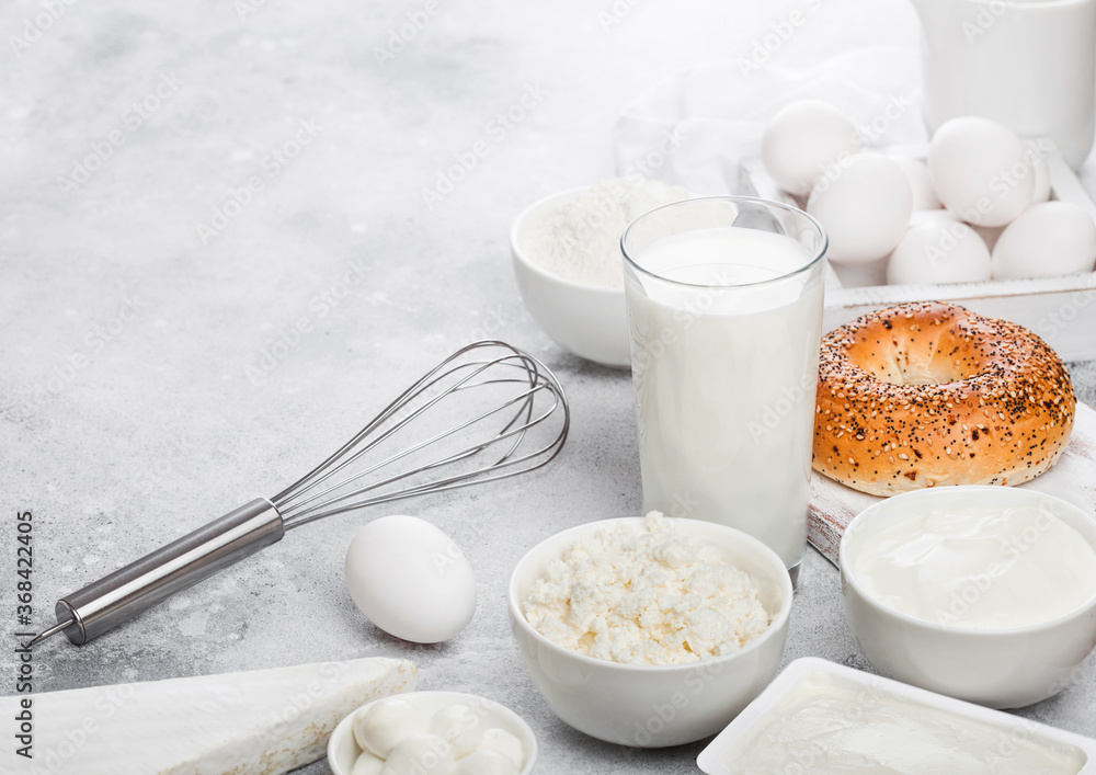 Fresh dairy products on white table background. Glass of milk, bowl of sour cream and cottage cheese and eggs in wooden box. Fresh baked baggel on round chopping board. Steel whisk. Space for text Fresh dairy products on white table background. Glass of milk, bowl of sour cream and cottage cheese and eggs in wooden box. Fresh baked baggel on round chopping board. Steel whisk. Space for text