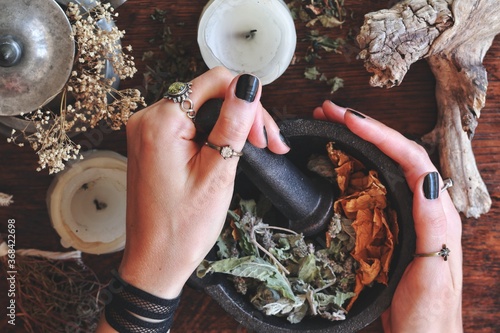 Kitchen witchery - female wiccan witch holding pestle and mortar in her hands, making magickal herb blend for a spell. Mix of dried herbs ready to be blended. Messy witch's altar on dark wooden table photo