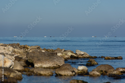 Stone sea shore and fishermen in boats on the horizon. Typical landscape of a seaside town. Fishing near the shore. Ultramarine blue of the sea and large stones. © Anna Pismenskova