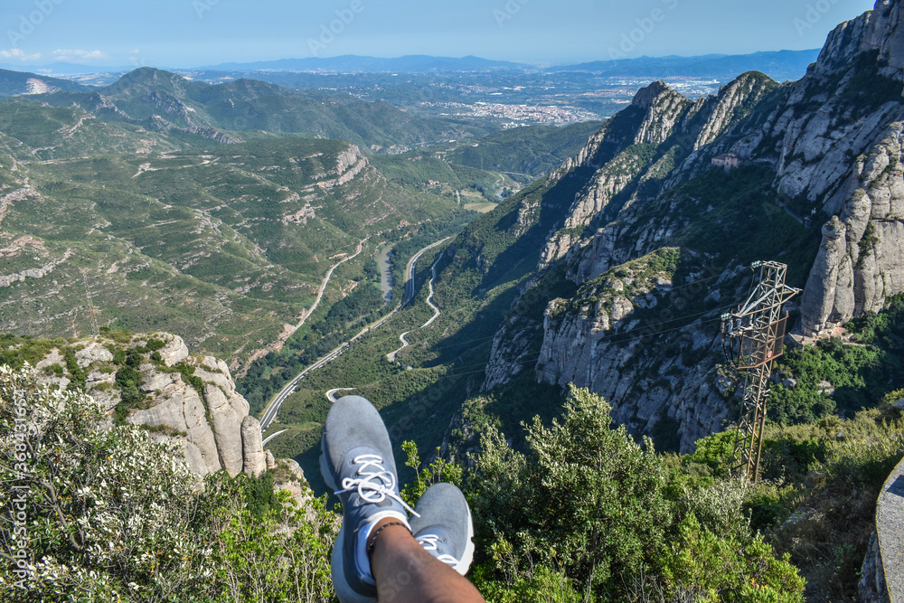 Man sitting with his legs dangling over a green valley,
 high mountains and a highway.