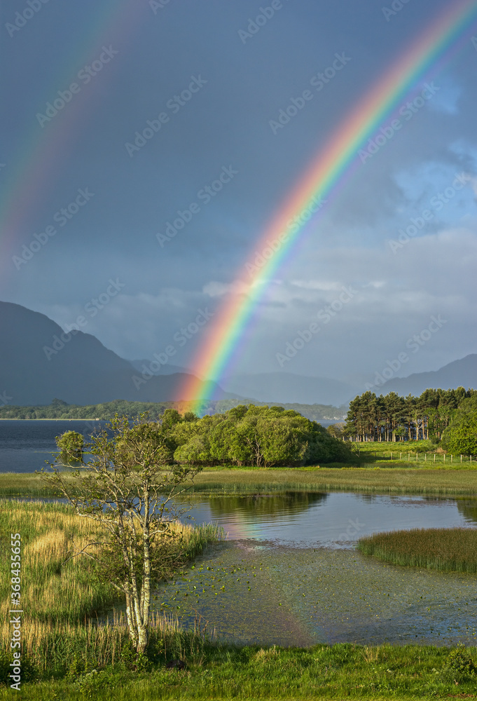 A genuine Irish double rainbow is reflected in the waters of Lough Leane, Killarney, Ireland, with the misty Kerry mountains in the background. Green bushes, reeds and rushes surround the lake shore. A genuine Irish double rainbow is reflected in the waters of Lough Leane, Killarney, Ireland, with the misty Kerry mountains in the background. Green bushes, reeds and rushes surround the lake shore.
