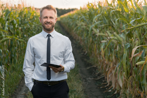Mature, well-dressed, businessman holding a new, black tablet, smiling and standing in the middle of green and yellow cornfield.