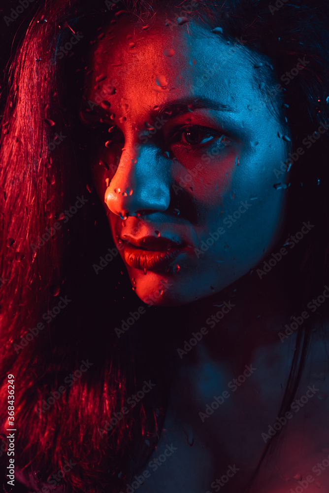 Portrait of sad young girl with red blue lighting behind glass with raindrops Portrait of sad young girl with red blue lighting behind glass with raindrops