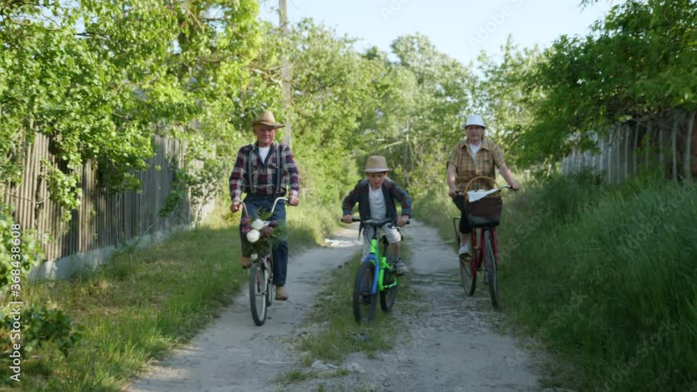 family holiday, happy male kid in sun hat with their merry grandmother and grandfather ride bicycles during outdoor activities in village family holiday, happy male kid in sun hat with their merry grandmother and grandfather ride bicycles during outdoor activities in village