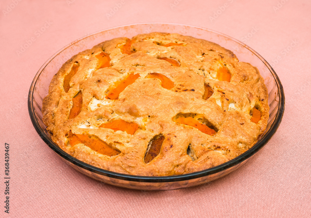 A large, round baked apple and apricot pie stands in a glass form on a table with a pink tablecloth. Delicious, fresh dessert. A large, round baked apple and apricot pie stands in a glass form on a table with a pink tablecloth. Delicious, fresh dessert.