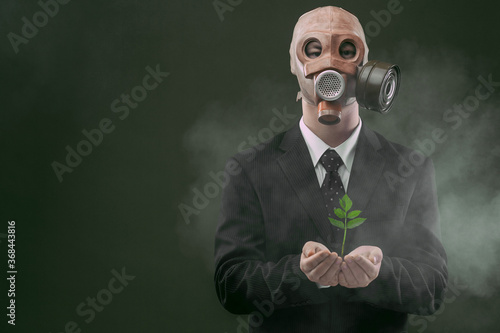 Portrait of a man a dark business suit and an old gas mask while holding a green plant, isolated on dark green background with fog. © Ion