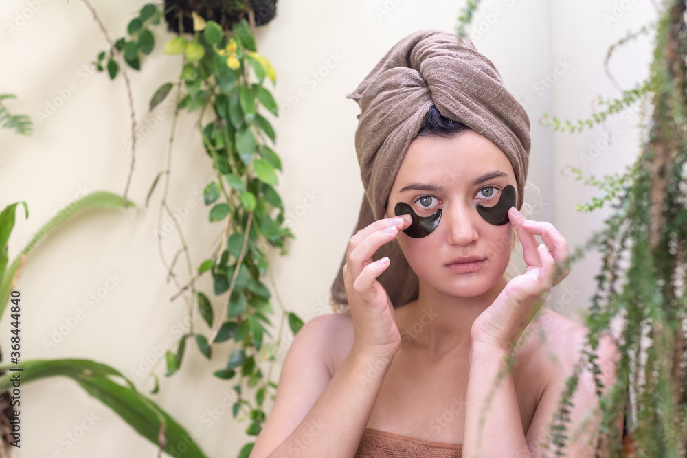 Young girl with a towel on her head applying hydrogel patches under her eyes in bathroom. Under eye masks for puffiness, lines, dark circles. Home skin care treatment Young girl with a towel on her head applying hydrogel patches under her eyes in bathroom. Under eye masks for puffiness, lines, dark circles. Home skin care treatment