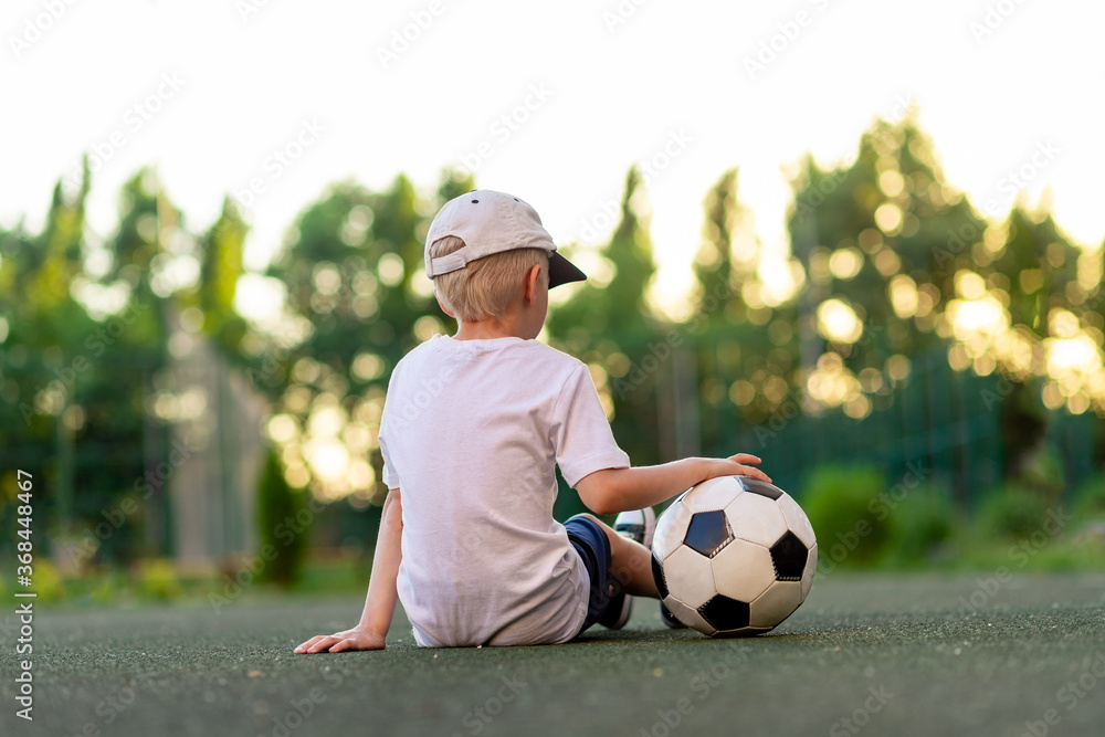 a boy in sports clothes sits on a green lawn on a football field with a soccer ball on his back at sunset, rear view, sports section, training