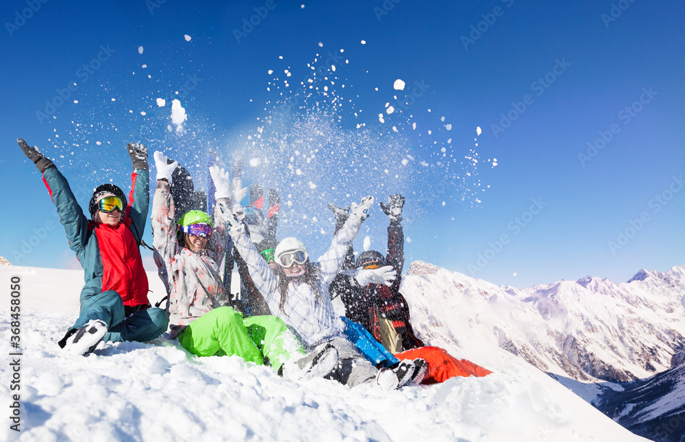 Group of young adults throw snow up in the air sitting on top of the mountain over sunny alpine peaks - obrazy, fototapety, plakaty 