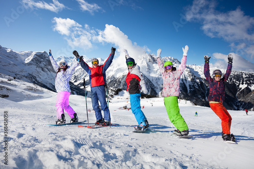 Group of young adults on ski and snowboards stand together in the row lifting hands over mountain tops