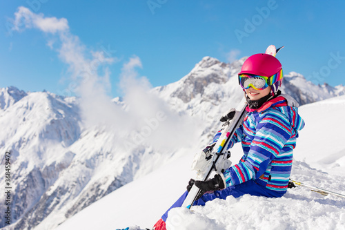 Happy young girl in bright sport outfit sit on the snow pile in the mountain over high peaks and clouds on sunny day