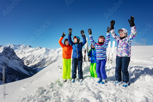 Big group of ski kids stand on snow rise hands cheerfully smiling over mountain range peaks in colorful sport outfit