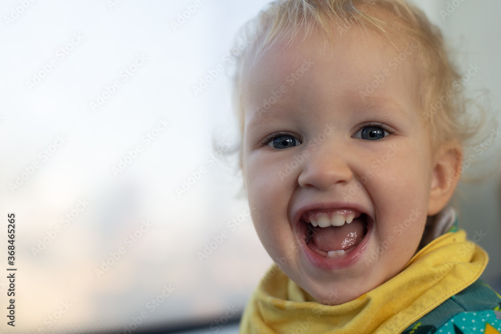 Happily laughing blond baby girl in front of window. Girl is wearing green and yellow clothes. Copy space available. Happily laughing blond baby girl in front of window. Girl is wearing green and yellow clothes. Copy space available.