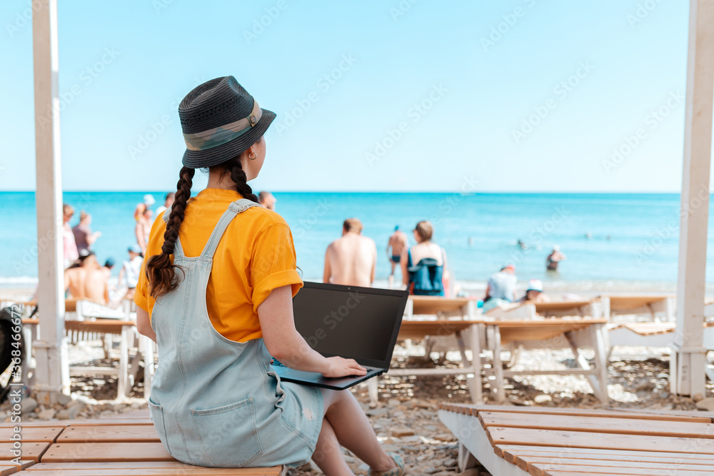 A young woman is sitting on a chaise longue and holding a laptop. Beach and sea in the background. Rear view. The concept of freelancing and vacation. Copy space