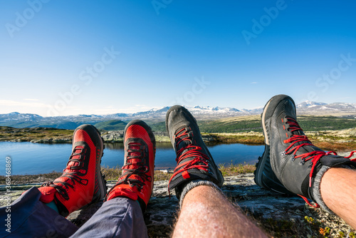 Couple with hiking boots on feet relaxing by lake during trek in the mountains during summertime.