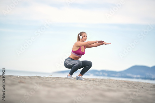.Girl listening to music and doing exercises on the beach.Woman squatting legs..