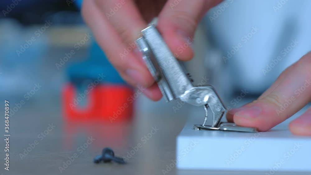 Carpenter man screws hinge to furniture door using electric screwdriver, hands closeup, side view. Carpenter is working making furniture in workshop. Doing renovation at home. Trying hinge on place. Carpenter man screws hinge to furniture door using electric screwdriver, hands closeup, side view. Carpenter is working making furniture in workshop. Doing renovation at home. Trying hinge on place.