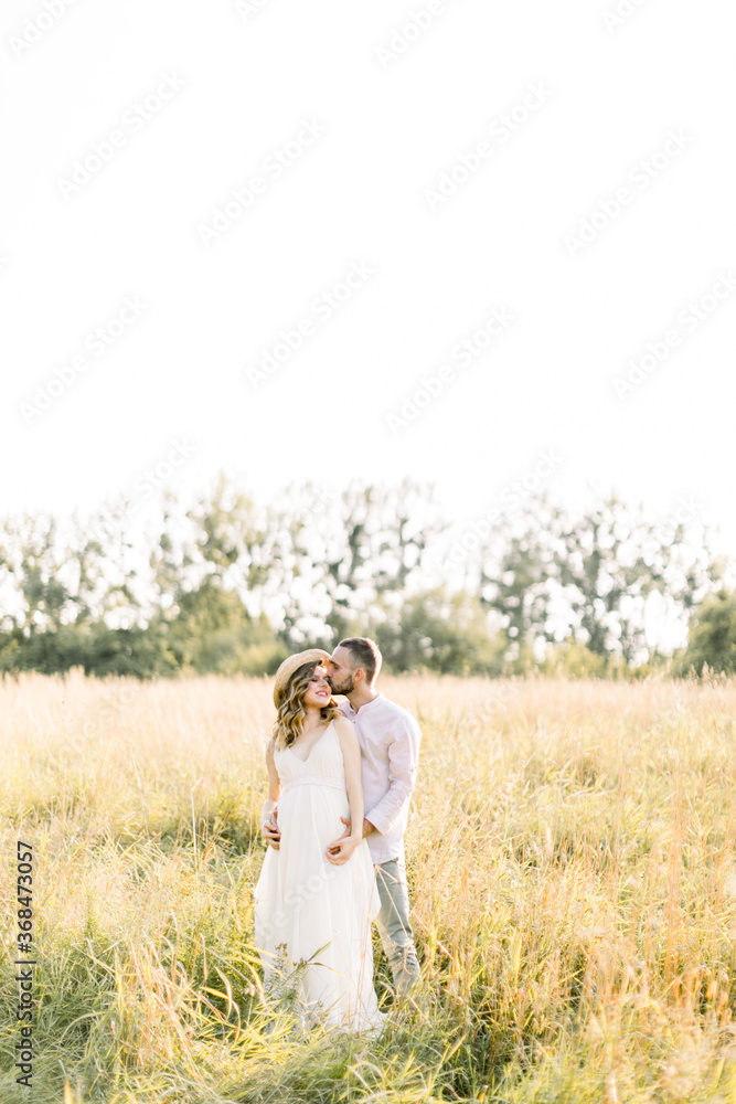 Young beautiful pregnant couple in white clothes walking in a field in the summer at sunset. Handsome bearded young man kissing his pretty pregnant lady in white dress and straw hat Young beautiful pregnant couple in white clothes walking in a field in the summer at sunset. Handsome bearded young man kissing his pretty pregnant lady in white dress and straw hat