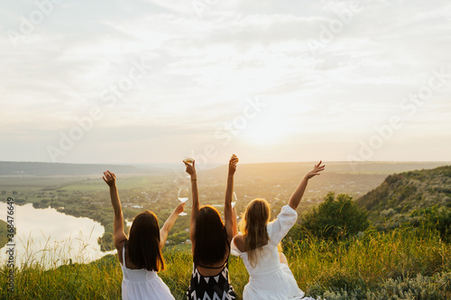 Three female friends sitting on the grass on the hill and having fun. View from behind. Young girlfriends having a picnic with river background. 