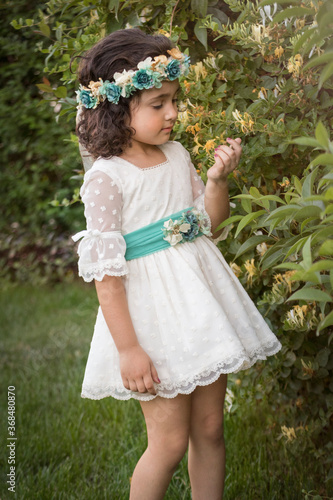 Girl in a white dress with green bow observing a flower in a garden. Curly brown hair.