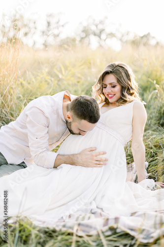 Pretty young pregnant woman with her handsome bearded man resting on a blanket in the summer meadow. Happy pregnant couple sitting on a blanket in a field