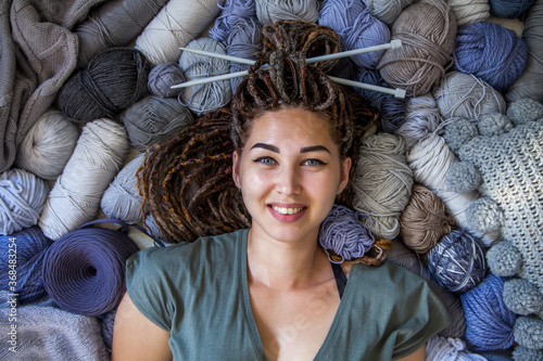 A beautiful young girl knitter lies and smiles surrounded by yarn for knitting. Female hobby and handicraft concept. Woman with dreadlocks. photo