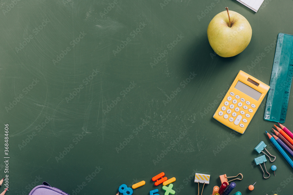 top view of ripe apple and calculator near school supplies on green chalkboard