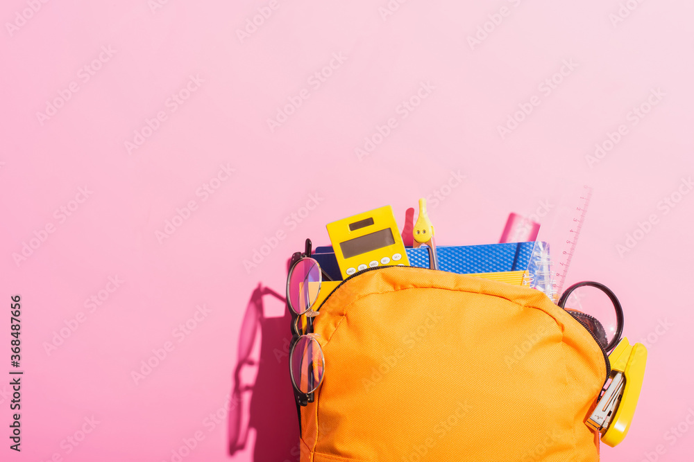 Yellow backpack packed with school stationery and eyeglasses on pink - obrazy, fototapety, plakaty 