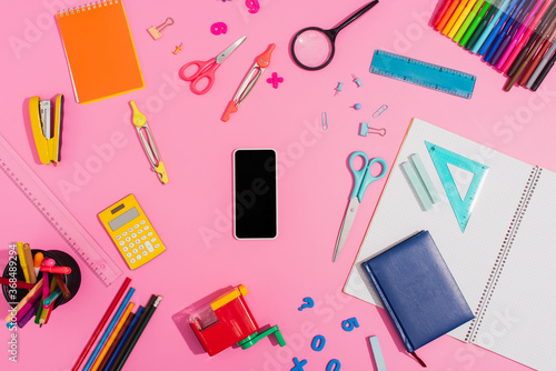 Top view of smartphone with blank screen surrounded by school supplies on pink