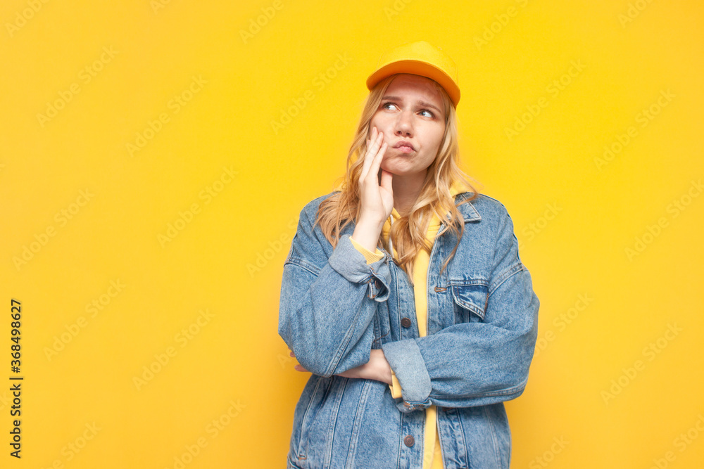 thoughtful girl in denim jacket and a cap stands on a yellow background and thinks, a hipster girl dreams and looks at a copy space thoughtful girl in denim jacket and a cap stands on a yellow background and thinks, a hipster girl dreams and looks at a copy space