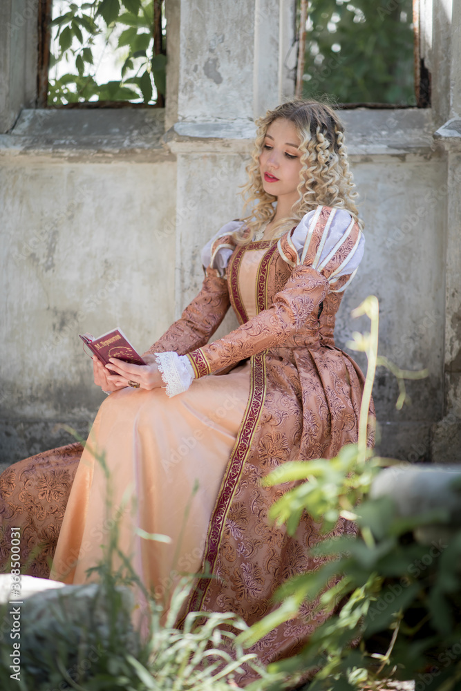 Young woman with white curly hair dressed in an medieval dress is sitting with a book near a castle