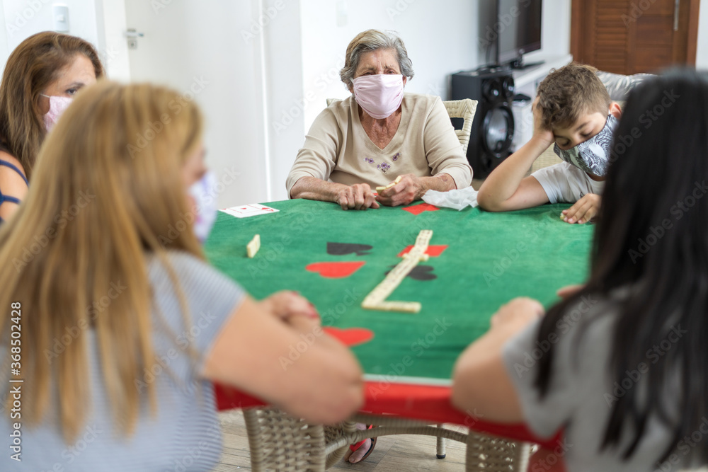 Fototapeta premium Família reunida dentro de casa durante quarentena, usando máscara de proteção, se divertindo jogando dominó e cartas.