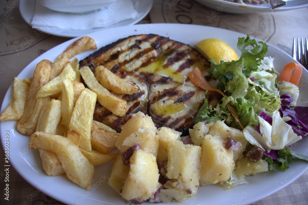 Food on a plate fish sword grilled french fries potatoes salad lemon olive oil in a cafe on the table Food on a plate fish sword grilled french fries potatoes salad lemon olive oil in a cafe on the table