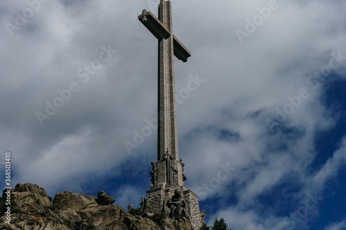 Cross on the top of the Valley of the Fallen in Madrid with white clouds in a very blue sky. The cross is settled on a rock hill