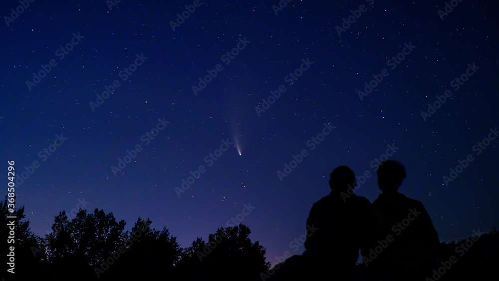 Silhouette of couple, man and woman looking at Comet NEOWISE in the night sky at sunset. Comet under the starry sky, in the evening, between the stars. Romantic family date near river and space.