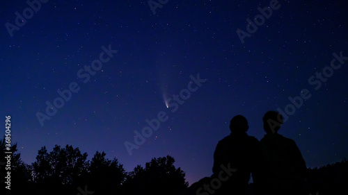 Silhouette of couple, man and woman looking at Comet NEOWISE in the night sky at sunset. Comet under the starry sky, in the evening, between the stars. Romantic family date near river and space.