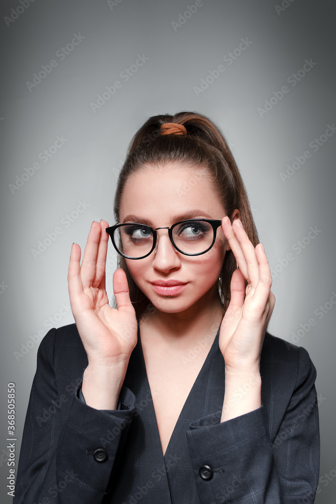 A young girl 20-25 years old in glasses, a jacket and with a tail in the image of a teacher poses on a gray background and shows different emotions
