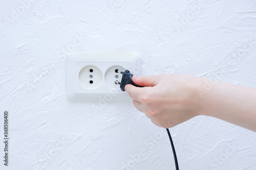 The hand of a Caucasian girl plugs the plug of an electrical appliance into white socket.