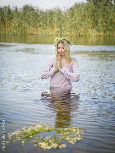 Young white woman with long blondy hair in a white dress in a river in summer with a wreath