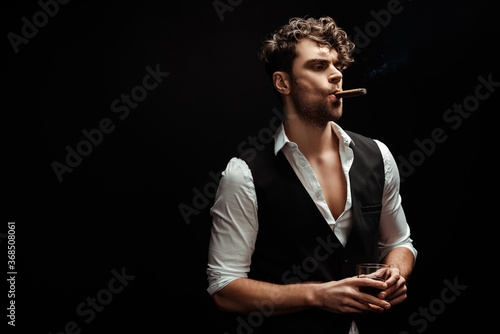 Fotografija Handsome man in shirt and waistcoat holding glass of whiskey and smoking cigar i