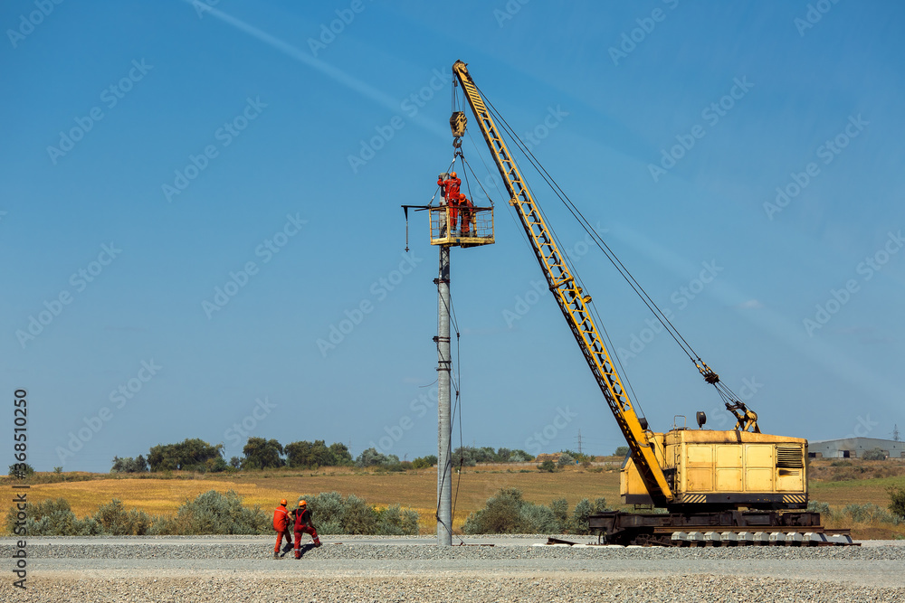 a railroad crane with a cradle lifted workers on the installation of electrical networks on a pole during the construction of a new railroad track, on sunny day and blue sky.