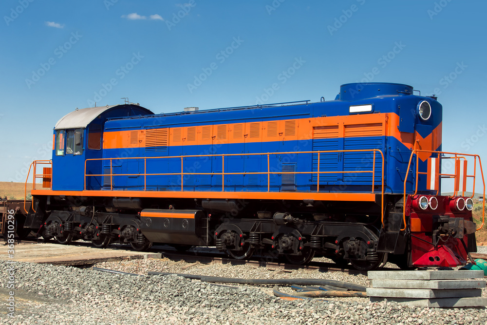 a blue shunting diesel locomotive with an orange stripe, a deck type locomotive stands on a railroad bed strewn with rubble, nobody.