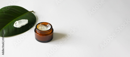 White beauty cream in brown glass jar and tropical leaf on background on white with copy space, horizontal banner format. Anti-aging moisturizer minimalistic. Zero-waste eco-friendly packaging photo