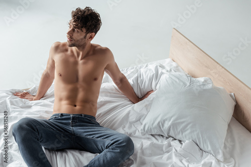 Muscular man in jeans looking away while sitting on bed on white background © LIGHTFIELD STUDIOS