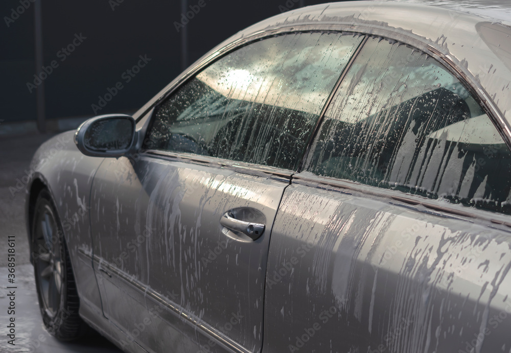 Outdoor car wash with soap. Fragment of gray vehicle with active foam. Sunny lighting. 
