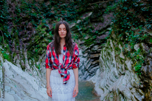 Young woman in white dress and plaid shirt practicing breathing yoga pranayama outdoors in moss gorge. Unity with nature concept.