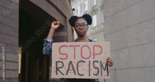 African american girl raising up clintched fist and holding carton cardbord with stop racism. Activist supporting equal human rights movement while standing at city street.Zoom in photo