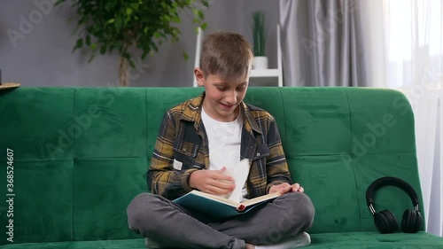 Front view of handsome joyful smart 10-aged boy which reading funny book at home on soft couch photo