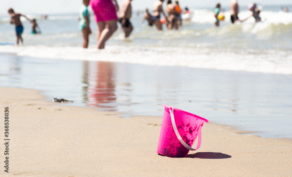 Plastic pink toy bucket left alone on the beach sand near water, blurred people on background - concept of vacation safety, drowning, safe summer playing for children.